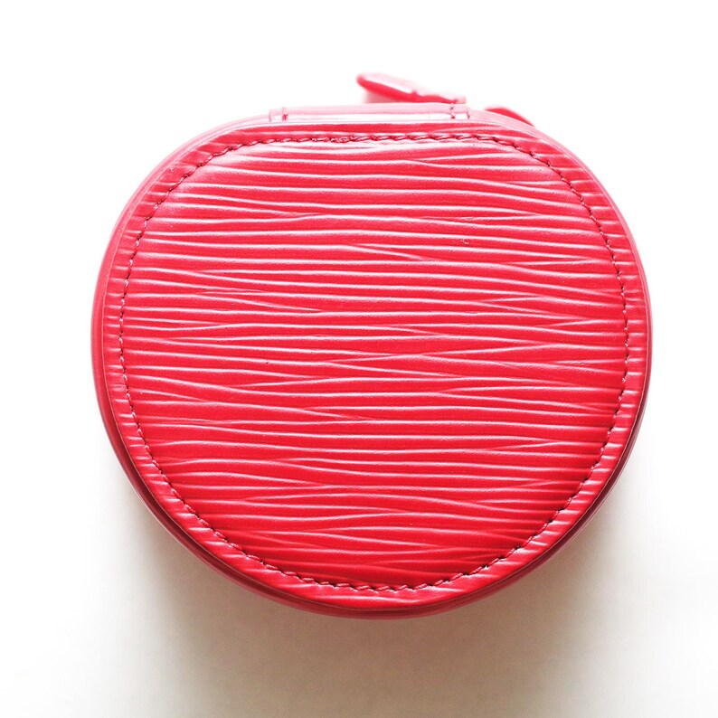 May include: A round, bright red jewelry case featuring a textured, horizontal wavy line pattern across its surface. The case is constructed with a stitched border around the circular lid and includes a small zipper pull at the top edge.