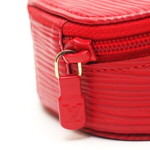 May include: A bright red textured leather case featuring a horizontal ribbed pattern. The case includes a matching red zipper closure with a gold-tone metal pull tab that displays an embossed LV logo on a red rectangular tag attached to the zipper.