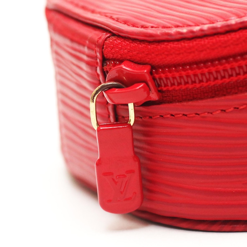 May include: A bright red textured leather case featuring a horizontal ribbed pattern. The case includes a matching red zipper closure with a gold-tone metal pull tab that displays an embossed LV logo on a red rectangular tag attached to the zipper.