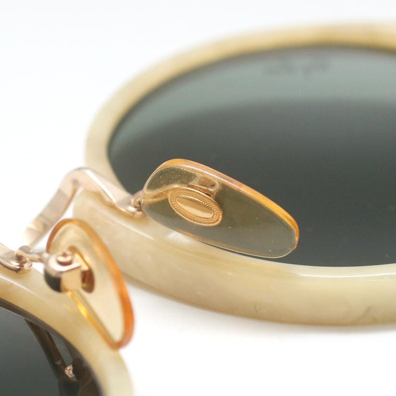 May include: Close-up of vintage sunglasses with a cream-colored frame and dark lenses. The nose pads and hinges are gold-toned. The sunglasses have a classic, round shape.