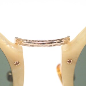 May include: Close-up of sunglasses with a gold-colored bridge and cream-colored frames. The bridge has a decorative dotted pattern. The lenses are a muted green color, and the frame is secured with small gold-colored screws.