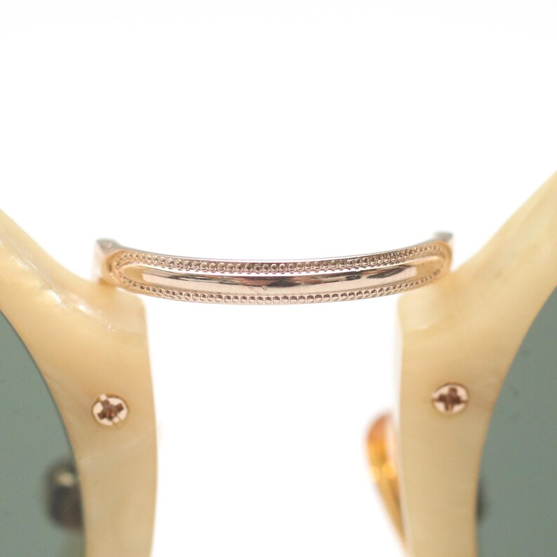 May include: Close-up of sunglasses with a gold-colored bridge and cream-colored frames. The bridge has a decorative dotted pattern. The lenses are a muted green color, and the frame is secured with small gold-colored screws.