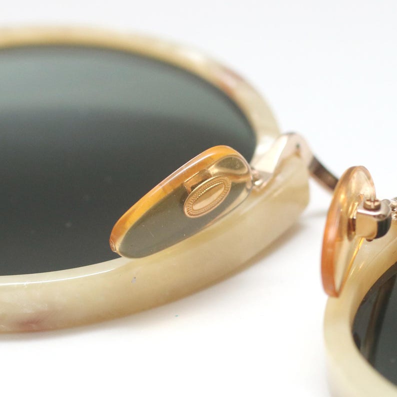 May include: Close-up of vintage sunglasses with a cream-colored frame and dark lenses. The nose pads are a translucent amber color, and the hinges are gold-toned. The sunglasses have a classic, round shape.