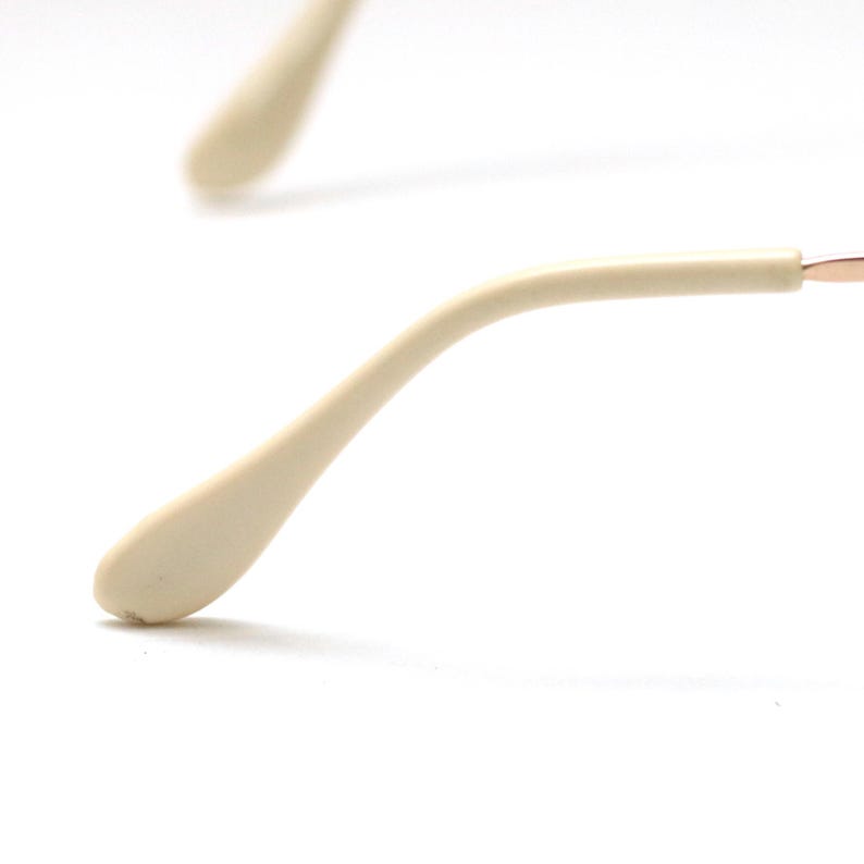 May include: Close-up of eyeglasses arm with a cream-colored temple and a thin, gold-colored frame. The temple curves gently, ending in a rounded tip. The background is plain white.