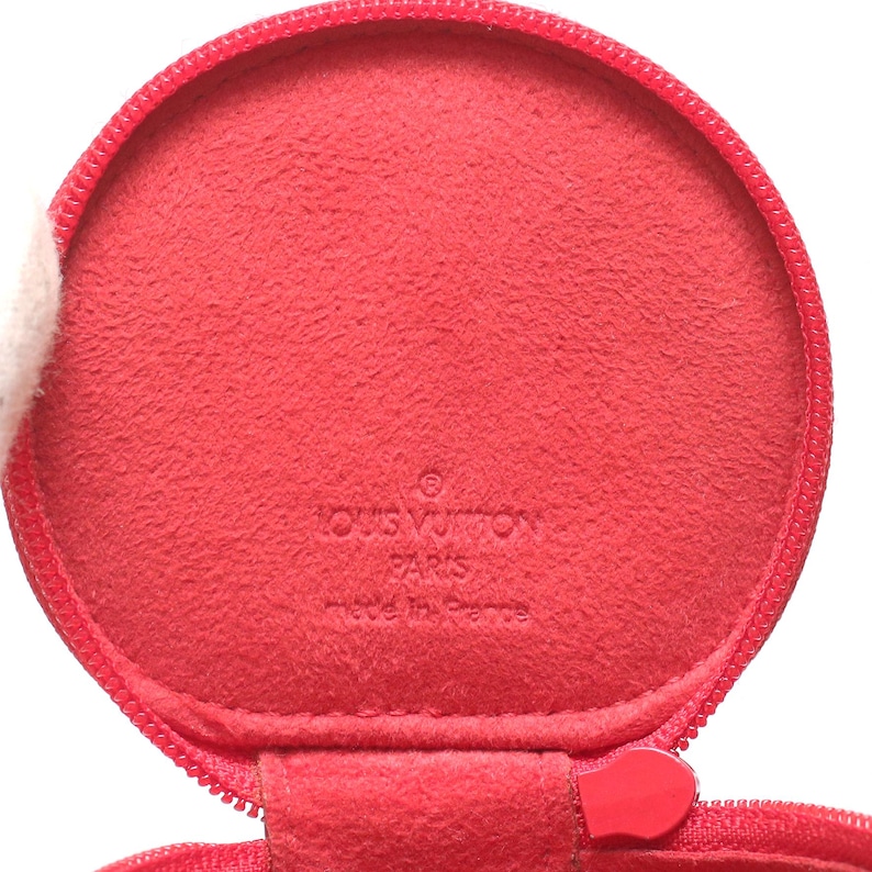 May include: A circular red coin purse with a zipper closure. The interior is lined with a matching red textured material. Embossed on the center of the interior is the text LOUIS VUITTON PARIS made in France, featuring a registered trademark symbol.