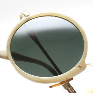 May include: Round, off-white sunglasses with dark green lenses. The frames have a marbled appearance and feature a small gold-colored accent on the top of the frame. The Ray-Ban logo is visible on the lens.