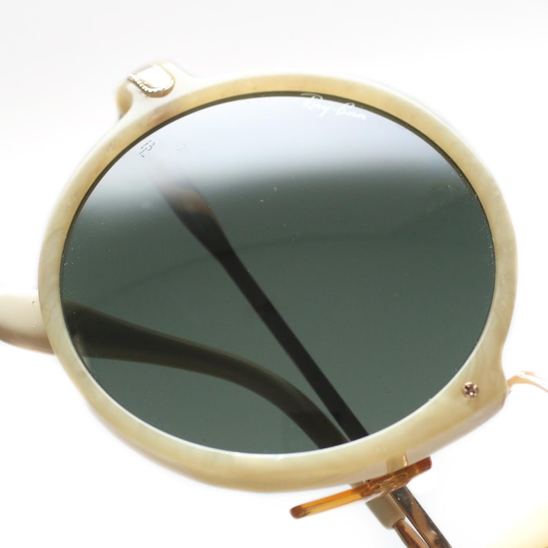 May include: Round, off-white sunglasses with dark green lenses. The frames have a marbled appearance and feature a small gold-colored accent on the top of the frame. The Ray-Ban logo is visible on the lens.