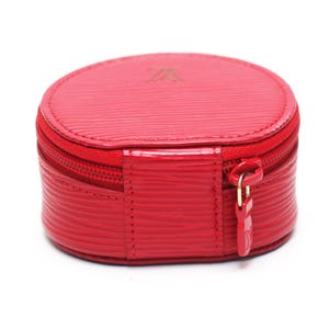 May include: A round, bright red jewelry case featuring a textured, horizontal-lined surface. The case has a zipper closure with a matching red pull tab embossed with a logo. A small, subtle logo is also embossed on the top center of the lid.