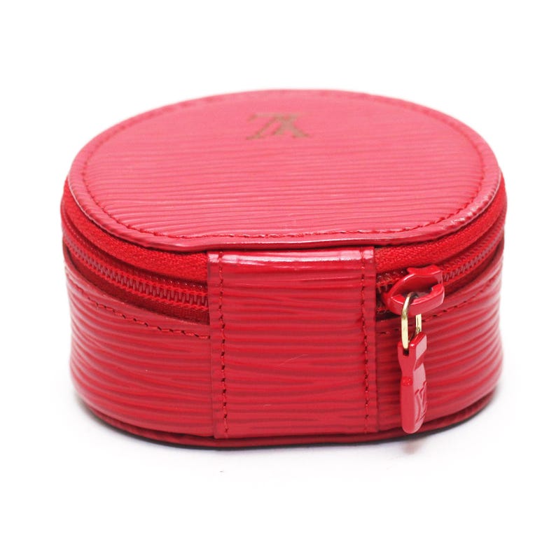 May include: A round, bright red jewelry case featuring a textured, horizontal-lined surface. The case has a zipper closure with a matching red pull tab embossed with a logo. A small, subtle logo is also embossed on the top center of the lid.
