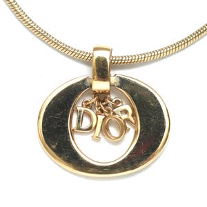 May include: A gold-toned necklace featuring a circular pendant with a smooth, polished finish. Suspended from the top of the circle are three individual gold-toned letters that spell out the word DIOR, hanging freely within the center opening.