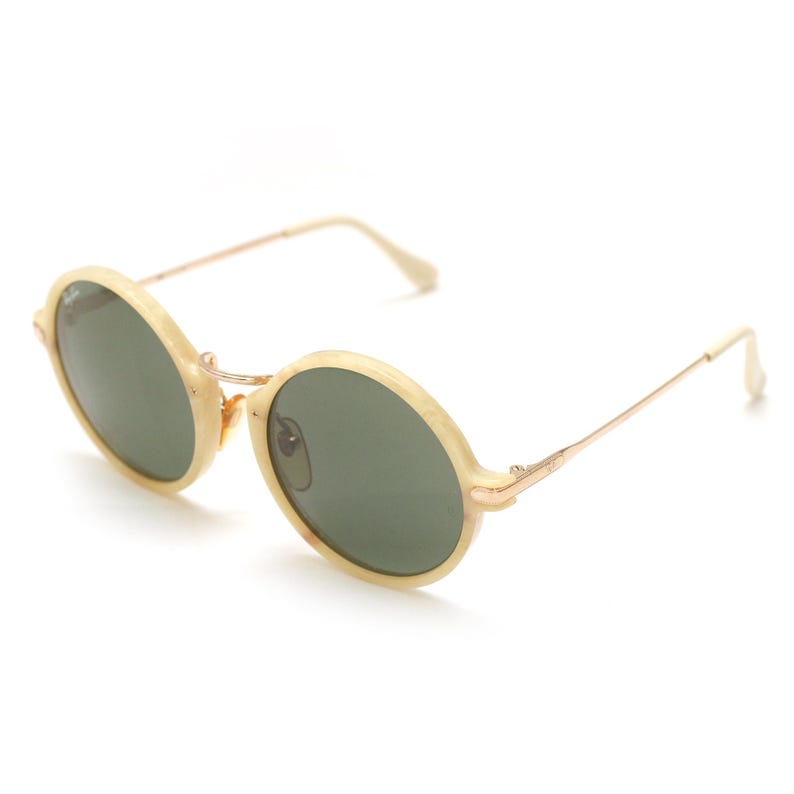 May include: Oval-shaped sunglasses with a light beige frame and dark green lenses. The frames are connected by a thin gold-colored bridge and arms. The sunglasses are angled slightly, showcasing their design.
