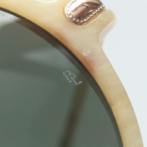 May include: Close-up of sunglasses with a light beige frame and dark green lenses. The frame has a gold-colored decorative element at the top corner. The lenses have the initials "B.L." etched on the surface.