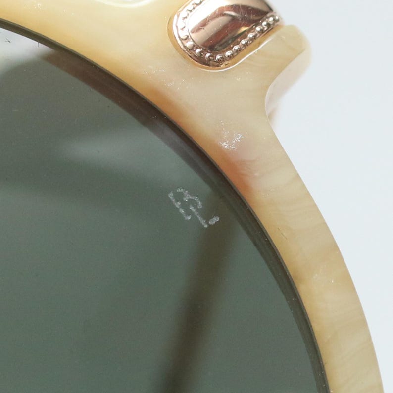 May include: Close-up of sunglasses with a light beige frame and dark green lenses. The frame has a gold-colored decorative element at the top corner. The lenses have the initials "B.L." etched on the surface.