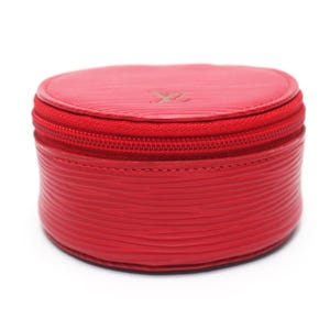 May include: A round, bright red jewelry case featuring a textured, horizontal ribbed surface. The case has a matching red zipper closure running around the circumference and a small, embossed LV logo centered on the top lid of the container.