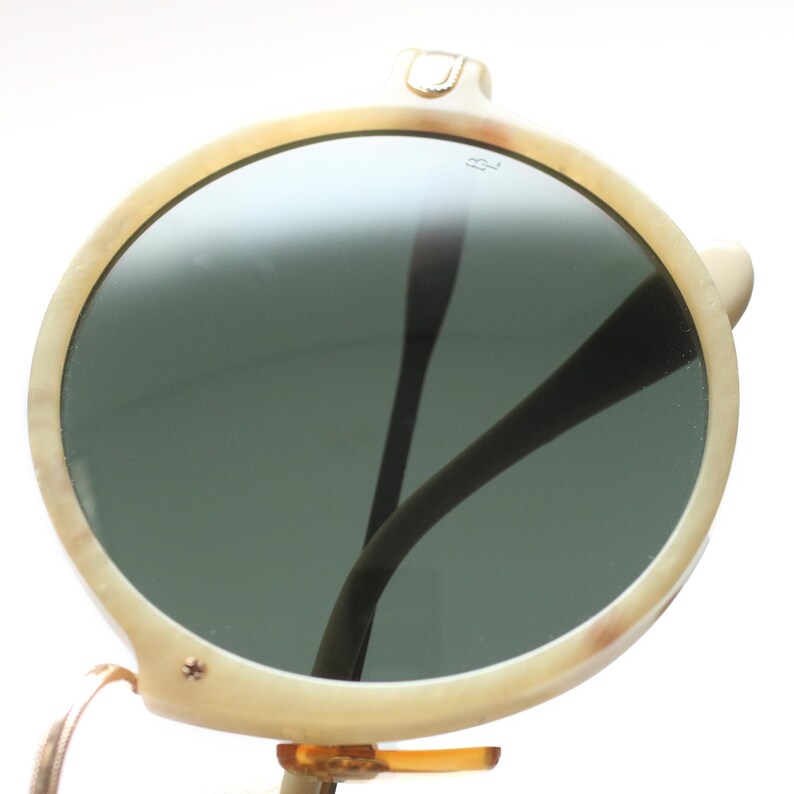 May include: Cream-colored round sunglasses with dark lenses. The frames have a marbled effect, and the temples are dark brown. The sunglasses have a small gold-colored accent on the top of the frame and a small gold-colored screw on the side.