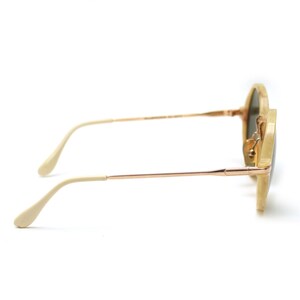 May include: Round sunglasses with a light beige frame and gold-colored metal arms. The lenses are a dark green color. The sunglasses are shown from a side profile view.