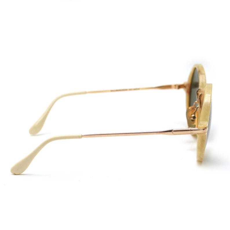 May include: Round sunglasses with a light beige frame and gold-colored metal arms. The lenses are a dark green color. The sunglasses are shown from a side profile view.