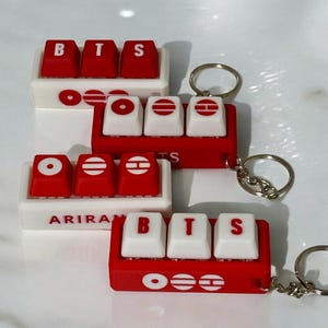 May include: Red and white keycap keychains with the letters "BTS" on them. The keychains have a red base and white keycaps, with silver key rings. The word "ARIRANG" is printed on one of the keychains.