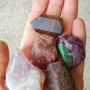 May include: A selection of polished gemstones of various colours and shapes are held in a hand. The stones include a dark brown, pointed crystal, a red and orange stone, a purple and green stone, a light purple stone, and a dark brown, rounded stone.