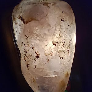 May include: A translucent, irregularly shaped stone with a light brown and white color palette. The stone has a rough, textured surface with dark brown inclusions. The background is a dark blue.