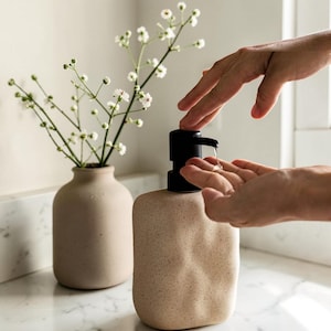 Puede incluir: Un dispensador de jabón de cerámica beige con una bomba negra. El dispensador tiene una superficie texturizada y una forma redondeada. Un pequeño jarrón con flores blancas está en el fondo. El entorno parece ser un baño.