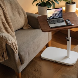 Pode incluir: Um laptop e um smartphone estão sobre uma mesa de madeira marrom. A mesa é suportada por um suporte branco ajustável com base retangular. Uma poltrona bege e um cobertor de malha estão ao fundo.