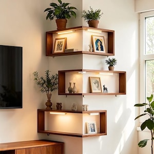 Puede incluir: Tres estanterías de esquina de madera con iluminación integrada, cada una exhibiendo obras de arte enmarcadas, pequeñas plantas en macetas de terracota y objetos decorativos. Las estanterías están montadas en una pared blanca, creando una estética moderna y minimalista.