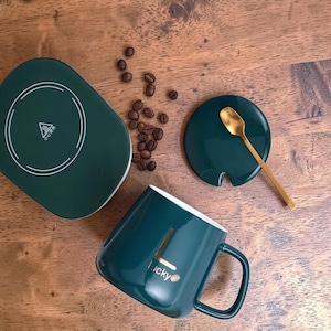 May include: A teal mug with the word "lucky" printed on it, a matching lid, and a gold spoon. A teal warming plate is also present. Coffee beans are scattered on the wooden surface.