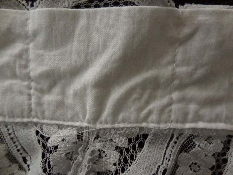 May include: Close-up of a white fabric with a lace trim. The fabric is folded and has a seam down the center.
