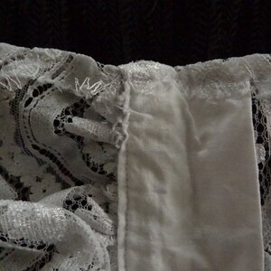 May include: White lace trim with a ruffled edge. The lace is delicate and has a floral pattern. The trim is attached to a white fabric.