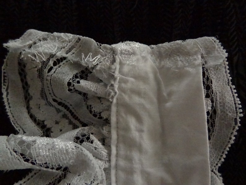 May include: White lace trim with a ruffled edge. The lace is delicate and has a floral pattern. The trim is attached to a white fabric.
