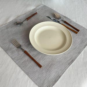 Op de afbeelding: Een crèmekleurige bord met een geribbelde rand ligt op een grijs-wit gestreepte placemat. Bestek met houten handvatten, waaronder een vork, lepel en mes, is rond het bord gerangschikt. De setting staat op een wit tafelkleed.