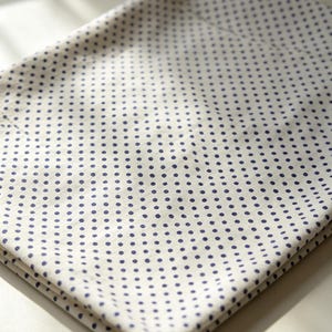 May include: A folded piece of fabric featuring a white base color with a repeating pattern of small, dark blue polka dots. The textile is arranged in a layered, rectangular fold, showcasing the consistent dot print across the visible surface area.