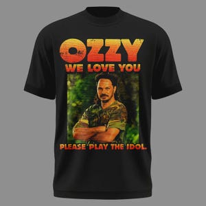 Peut inclure: T-shirt noir avec le mot "OZZY" en orange et jaune, et le texte "WE LOVE YOU". En dessous, une image d'un homme en chemise camouflage, avec le texte "PLEASE PLAY THE IDOL" en dessous.