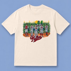 May include: A cream-colored short-sleeve t-shirt featuring a graphic of five baseball players and a green mascot standing in a cornfield. Below the illustration, the word Phillies is printed in a red, cursive, script-style font.