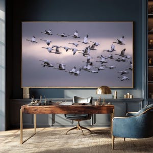 May include: A large framed artwork depicts a flock of white birds in flight against a dusky sky. The artwork hangs above a wooden desk with a gold-toned lamp and a blue armchair. The room has dark blue walls and a patterned rug.