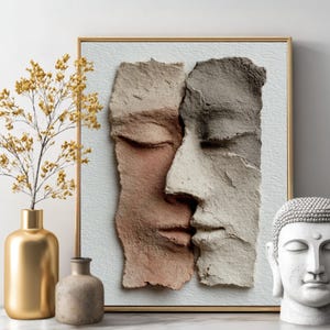 Puede incluir: Una obra de arte enmarcada con dos caras esculpidas, una rosa y otra gris, una al lado de la otra. Un jarrón dorado con ramitas amarillas y un jarrón pequeño de tonos neutros a la izquierda. Una estatua de cabeza de Buda blanca a la derecha.