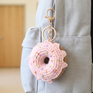 以下が含まれることがあります： 薄いピンク色のアイシングとカラフルな刺繍のトッピングが施された、かぎ針編みのドーナツ型キーホルダー。ドーナツは金色の金属製ナスカンと二重リングに取り付けられており、ライトグレーの布製バックパックにぶら下がっています。