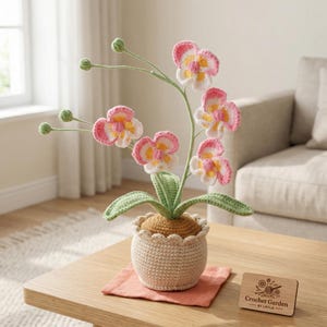 May include: A crocheted orchid plant with pink, white, and yellow flowers in a beige pot. The plant sits on a small, square, peach-coloured cloth. A wooden sign with the text "Crochet Garden by Layla" is next to the plant.