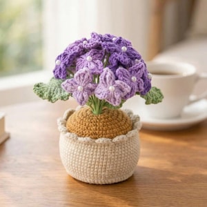 Puede incluir: Una planta de hortensia de ganchillo en una maceta beige. Las flores moradas tienen centros de cuentas blancas y las hojas verdes añaden un toque natural. La maceta tiene un borde festoneado y se asienta sobre una superficie de madera.