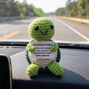 May include: A green and white crocheted turtle figurine holding a sign that reads "Positive Turtle." The sign says "Tiny turtle is always here whenever you need a little cheer. YOU'RE AWESOME!" The turtle is sitting on a car dashboard.