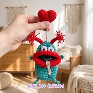 May include: A handmade crochet key holder in the shape of a whimsical monster. The teal body has red lips, eyes, and hair-like accents. A red heart is attached to the top. The text "Keys not included" is at the bottom.