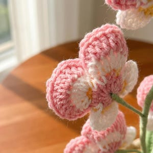 May include: Close-up of a handmade crochet flower with pink and white petals. The flower has a detailed, textured appearance, with visible stitch patterns. The stem is green. The background is blurred, suggesting a soft focus.