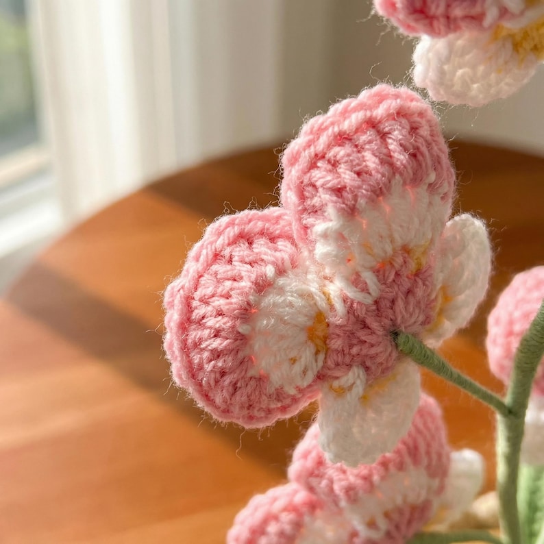 May include: Close-up of a handmade crochet flower with pink and white petals. The flower has a detailed, textured appearance, with visible stitch patterns. The stem is green. The background is blurred, suggesting a soft focus.