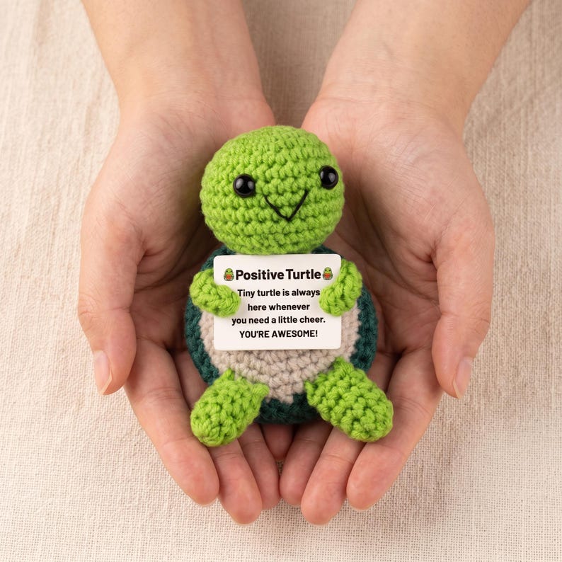 May include: A handmade green crochet turtle with a smiling face and a white sign that reads "Positive Turtle". The sign says "Tiny turtle is always here whenever you need a little cheer. YOU'RE AWESOME!"