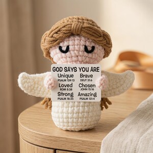 May include: A handmade crochet angel doll with a light brown halo and hair, holding a sign that reads "GOD SAYS YOU ARE Unique, Brave, Loved, Chosen, Strong, Amazing" with Bible verses. The angel has white wings and is sitting on a wooden surface.