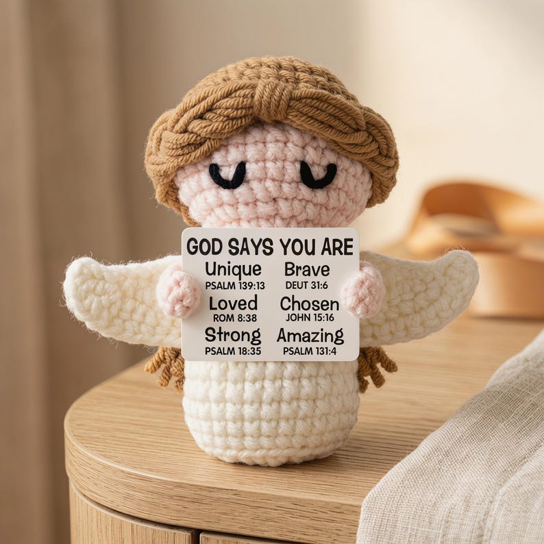 May include: A handmade crochet angel doll with a light brown halo and hair, holding a sign that reads "GOD SAYS YOU ARE Unique, Brave, Loved, Chosen, Strong, Amazing" with Bible verses. The angel has white wings and is sitting on a wooden surface.