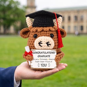 May include: A brown crocheted highland cow plush wearing a black graduation cap with a red tassel. The cow holds a white scroll with a red ribbon and a rectangular sign that reads: CONGRATULATIONS! GRADUATE So very Extremely Amazing Super duper Proud of YOU!