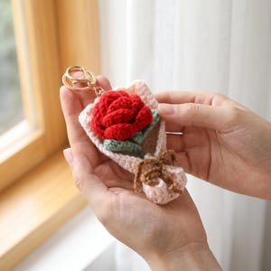 May include: A crochet keychain featuring a red rose with green leaves, wrapped in a light pink crocheted bouquet holder tied with a brown yarn bow. The accessory includes a gold-toned metal lobster clasp and a circular split ring for attachment.