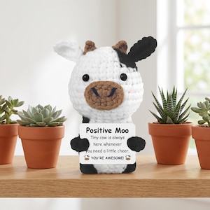 Op de afbeelding: Een gehaakte koeienfiguur met een zwart-wit patroon, met een bordje met de tekst "Positive Moo". Op het bord staat ook "Tiny cow is always here whenever you need a little cheer. YOU'RE AWESOME!" De koe is omringd door kleine potplanten.